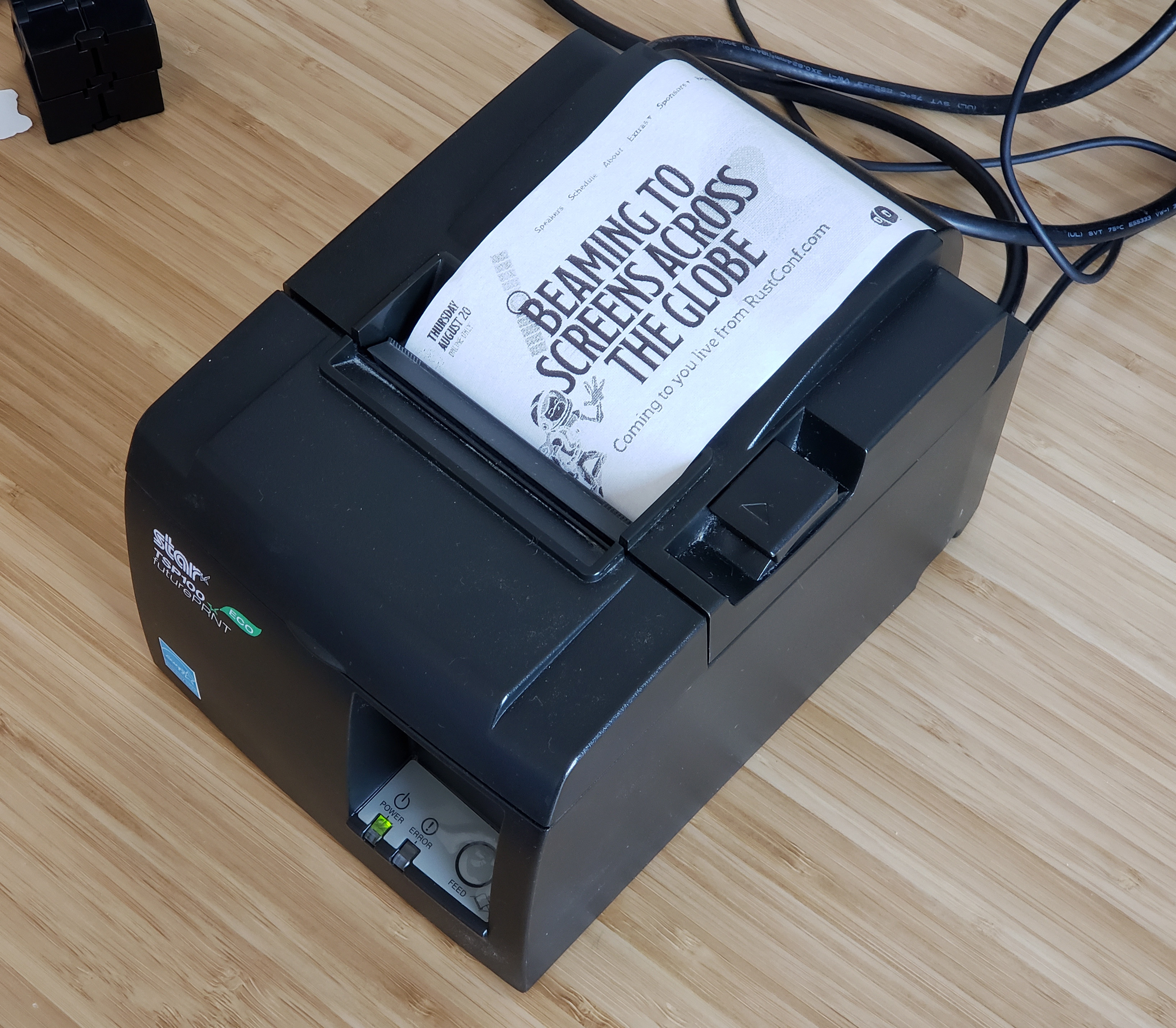 A Star TSP100 Eco futurePRNT 72mm receipt printer, powered on with a printed receipt showing the RustConf homepage reading "Beaming to screens across the globe"